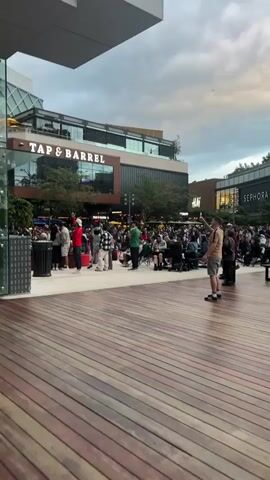 The video shows an outdoor plaza or walkway with wooden planks. In the background, there is a building with the sign "TAP & BARREL." Further in the background, there are other stores, including "H&M" and "SEPHORA." A crowd of people are gathered in front of the "TAP & BARREL" establishment. Some are standing, while others are seated at tables. The sky is overcast. In the foreground, a man in a tan shirt and shorts is walking and looking at his phone. Another person in a black shirt and jeans is standing to his right. The overall atmosphere appears to be that of a public gathering or event.