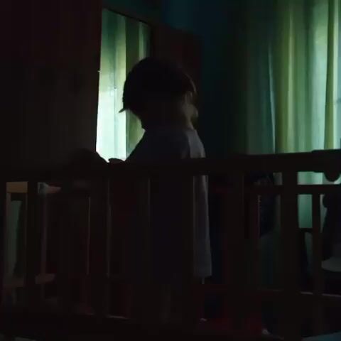 A child is standing in a crib. The child is wearing a light-colored t-shirt and dark shorts. The child is facing away from the camera, towards a window with green curtains. The child is holding a doll. The crib is made of wood. There is a wardrobe in the background. The lighting is dim.