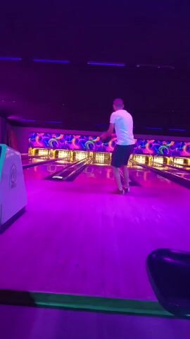 A man in a white t-shirt and black shorts bowls in a dimly lit bowling alley. The lanes and walls are illuminated by vibrant neon lights, creating a purple and pink glow. The man releases the bowling ball and watches it roll down the lane. He then turns and walks back towards the camera, looking at the viewer.