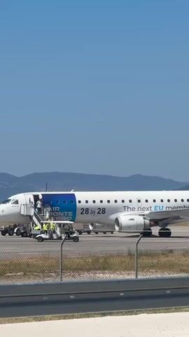 The video shows an airplane with "AIR MONTE NEGRO" written on its side, parked on an airport tarmac under a clear blue sky. Several people are visible near the plane's entrance stairs. Some appear to be passengers disembarking, while others, wearing high-visibility vests, might be airport staff assisting with the process. A baggage cart is also present on the tarmac. In the background, hills are visible against the horizon. The scene appears to be a normal airport operation.