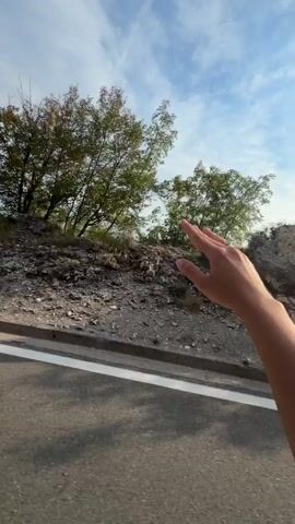The video shows a person's right hand and forearm extending out of a vehicle window. The hand is held up, fingers splayed, as if feeling the air or gesturing. The vehicle appears to be moving, indicated by the blurred road surface in the foreground and the passing scenery. The background consists of a rocky, vegetated hillside with trees, under a partly cloudy sky. Beyond the immediate hillside, a vista of rolling mountains and a valley unfolds. The video progresses through a series of slightly different camera angles and slight movements, but the core action remains the same: a hand outside a moving vehicle.