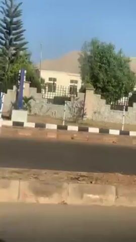 The video depicts a street scene with a building on the opposite side of the road. The building is beige with a light-colored roof and features a stone fence with black iron bars. The road is paved and has a black and white curb. An auto rickshaw passes by on the road. The sky is clear and blue. The ground along the roadside is dirt-colored and appears somewhat eroded. The perspective is that of someone filming from a moving vehicle.
