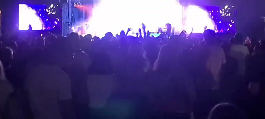 This is a low-resolution, shaky, night-time video of a live concert. The stage is brightly lit with white spotlights. There are large screens on either side of the stage displaying abstract, sparkling blue imagery. The crowd in the foreground is mostly silhouetted and appears to be enjoying the performance. Several musicians are visible on stage, some with instruments. The overall atmosphere is energetic and festive.