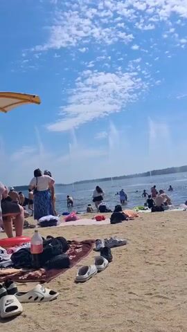 This is a medium shot of a crowded beach on a sunny day. The sky is blue with scattered white clouds. Several people are in the water, some of whom appear to be swimming or wading. Others are on the sand, sitting or standing. There are towels and various personal items spread out on the beach. In the background, several water fountains are visible in the lake. An umbrella is partially visible in the top left corner of the frame.