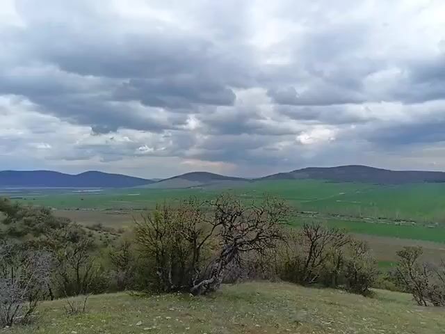The video shows a landscape with green fields and hills under a cloudy sky. In the foreground there are some trees and grass. The camera slowly pans from left to right, revealing more of the valley and surrounding hills. The sky is predominantly grey but with some patches of blue.