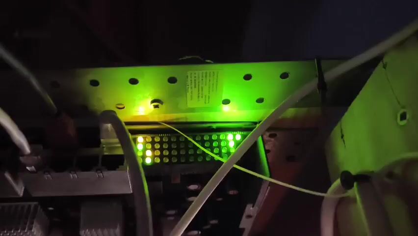 The video shows a close-up view of electronic equipment with various green and yellow lights. The perspective is from below, looking up at a panel with rows of small lights that are illuminated. Some cables are visible, running across the equipment. The lighting suggests the environment is dimly lit.