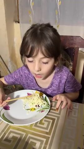 A young girl with dark hair and a purple shirt is sitting at a table and eating noodles with chopsticks. She is looking at the camera and her mouth is open as she eats. The table has a geometric pattern on it, and there is a plate with noodles in front of the girl. There is also a chair behind her. The background is blurred but appears to be an indoor setting with curtains.