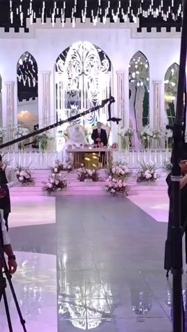 The video shows an indoor event, likely a wedding reception or formal party. The venue is elaborately decorated with white flowers and lights hanging from the ceiling, creating a canopy effect. White pillars with green accents line the sides of the room, and a long, illuminated aisle runs down the center. Several people are present, including guests seated at tables and crew members operating video equipment. In the foreground, a DJ or sound technician is positioned behind a soundboard. Further down the aisle, three cameramen are actively filming, two with tripods and one with a handheld camera. At the end of the aisle, a couple is standing at a decorated altar or stage, presumably the focus of the event. The overall atmosphere appears festive and formal.