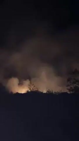 Here's a description of the video:

The video depicts a fire at night. The flames are visible behind a line of trees, and there is smoke rising into the air. There are visible sparks of light coming from within the fire. The environment appears dark and the only light source is the fire itself, which illuminates the smoke and the nearby trees. The overall impression is one of a potentially serious uncontrolled fire in a wooded area.
