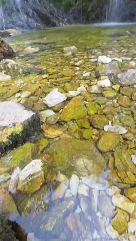 The video shows a view of a waterfall from the bottom, looking up towards the cascading water and the moss-covered rocks behind it. The water is clear, and the bottom of the pool is filled with smooth, multicolored stones. The scene appears to be natural and serene.