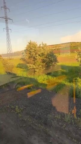 The video shows a view from a moving vehicle, likely a train or bus, looking out a window. The landscape is rural, with a large power line tower dominating the left side of the frame. The ground in the foreground is covered with dark gravel and some sparse green vegetation. Beyond that, a field of tall, green grass stretches towards a gently rolling hill in the distance. The field is dotted with small yellow and red flowers, and a few scattered trees. The sky is clear and blue, with the warm light of late afternoon or early morning casting long shadows across the field. There are reflections on the window, obscuring some of the finer details of the scene. No people are visible.
