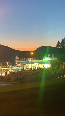 The video shows an outdoor swimming pool complex at dusk. The sky is a deep blue with an orange and pink gradient near the horizon. In the background, a large, multi-story building resembling a cruise ship is illuminated with colorful lights, featuring windows, decks, and a funnel-like structure. Several swimming pools are visible, filled with people. There are also lounge chairs and umbrellas scattered around the pools, and food stands or kiosks are present. Trees and hills form the backdrop to the scene, and a tall lamp post with a bright light stands in the foreground. People are seen walking, sitting, and swimming in the pools, suggesting a lively evening atmosphere at the venue.