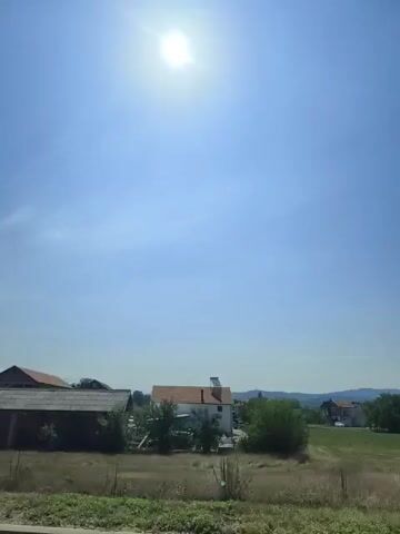 The video pans across a rural landscape under a bright, sunny sky. In the foreground, dry grass and a wire fence are visible. In the midground, several houses and outbuildings are scattered across the scene, some appearing to be under construction. Trees and bushes are interspersed among the structures. In the background, hills can be seen in the distance. The sun is high in the sky, creating a glare.