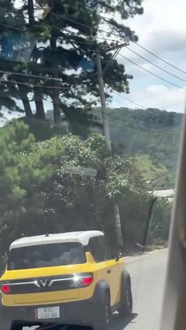 A yellow VinFast VF 3 electric car is driving on a paved road through a mountainous, wooded area. The car has a white roof and black wheel wells. Its license plate reads "81C 230.96". The road curves to the left. In the background, there are green hills with scattered trees and some areas with exposed red earth and black netting, possibly indicating agriculture or construction. Power lines are visible overhead. The sky is cloudy.