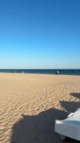 This media file captures a serene beach scene, likely during a warm, sunny day given the clear blue sky and bright sandy expanse. The foreground is dominated by a stack of white plastic lounge chairs, casting long shadows across the textured sand, suggesting it is late afternoon or early morning. Beyond the chairs, the beach stretches towards the ocean, dotted with a few people engaged in various leisurely activities. Some individuals are standing near the water's edge, while others are further out in the calm, deep blue sea. The horizon is marked by the distinct line where the sea meets the sky, with a few distant buoys or markers visible on the water. A cluster of straw umbrellas and more lounge chairs are visible to the right, hinting at a developed beach resort or public area. The overall atmosphere is tranquil and inviting, evoking a sense of relaxation and summer vacation. The location context provided is Lagos, Portugal, which aligns with the typical coastal scenery of the region. No visible text is present in the media.