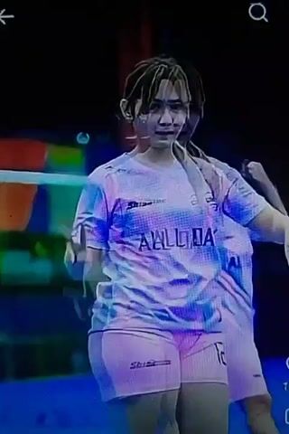 The video shows two young women in athletic attire on what appears to be a sports court. The first woman, in the foreground, is wearing a tie-dye style shirt with pink and blue swirls and the words "ALL DAY" printed on it, along with pink athletic shorts. She is facing the camera and holding up two fingers in a peace sign. Her hair is pulled back in a ponytail. Behind her, to the right, a second woman in a similar pink and tie-dye athletic outfit is visible from the side. In the latter half of the video, the focus shifts to the second woman, who is now in the foreground. She is wearing a white athletic shirt with red sleeves and a pattern of small red flowers, along with red shorts and knee pads. She is running and looking forward. The background shows stadium seating with dark and colorful sections.