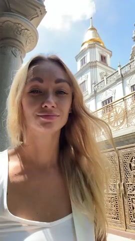The video shows a woman with long blonde hair standing in front of a temple complex, taking a selfie. The woman wears a white top, and her hair is slightly windswept. The temple buildings are white with ornate carvings and gold-colored spires. The sky is blue and sunny. As the video continues, the woman rotates to show more of the temple grounds, including trees and a stone pathway.