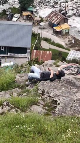 The video shows a group of people resting on rocks near a river. In the foreground, a person lies on their back on a rocky outcrop, dressed in a black shirt and blue jeans, wearing sunglasses and a hat. Further back on the rocks, a group of people are seated or lying down. One person, wearing a red and blue striped shirt and a dark cap, is looking down at their hands. Another person, wearing a patterned scarf and a dark top, is lying on their side. Several bags and blankets are scattered on the rocks. In the background, a river flows through a rocky terrain. Several small buildings with corrugated roofs are visible near the river, along with some vehicles. The landscape is mountainous, with green hills and trees, and the sky is cloudy. The overall impression is of a resting spot in a natural, somewhat rugged environment.