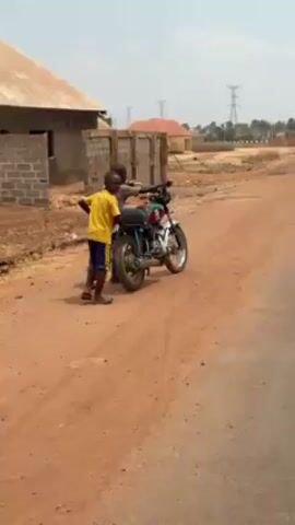 Two children are pushing a motorcycle down a dirt road. The child in the back is wearing a yellow shirt and blue shorts. The child in front is sitting on the motorcycle. The motorcycle has a custom paint job. There are several houses in the background, some of which appear to be under construction. The road is dusty and red. The sky is overcast.
