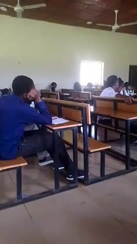 The video shows the interior of a classroom from a low angle, focusing on rows of empty wooden desks with black metal frames. Several students are seated at the desks. In the foreground, a student wearing a blue shirt and dark pants is seated facing left, with their head resting on their hand. Further back, other students are visible, some appearing to be paying attention, while others seem disengaged. A whiteboard is present at the front of the room, with a person standing in front of it, possibly a teacher or lecturer, writing on the board. The room has a tiled ceiling with two ceiling fans and a window on the left side letting in natural light. The overall atmosphere appears to be a typical lecture or class setting.