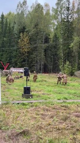 This is a video taken from ground level, looking towards a grassy field with a line of trees in the background. In the foreground, tall, dry grass is visible. In the middle ground, several people in historical military uniforms are present. Some are crouching or kneeling, while others are standing. One person is standing near a black speaker on a stand. Two individuals in uniform are facing each other, one holding what appears to be a rifle. Further to the left, another group of people in uniform are gathered. A red object, possibly part of a prop or structure, is visible on the far left. In the background, a green, tank-like structure is partially visible on the right. Power lines are visible stretching across the trees. The overall scene suggests a historical reenactment.