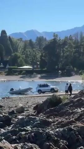 The scene shows a calm lake on a sunny day with a white SUV parked on a ramp partially submerged in the water, towing a small inflatable boat. Two people are walking away from the water's edge on a rocky shoreline. Behind the vehicle, there is a sandy beach lined with trees and buildings, including a white structure and a red building. In the background, mountains can be seen. The foreground is dominated by rocky terrain.