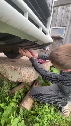 A young man with short brown hair, wearing a grey hooded jacket with black stripes and grey sweatpants with black stripes, is kneeling on the ground. He is holding a silver tool, possibly a screwdriver, and appears to be working on the underside of a white vehicle. The vehicle is propped up on large, rough-hewn grey stones and several logs, suggesting it is being raised for maintenance. Green foliage and grass surround the area. In the background, a wooden structure, possibly a shed or barn, is visible, with a small framed object hanging on its wall. A red and black power tool, likely a drill, is also present on the ground near the vehicle. The man is looking towards the camera with a slight smile.