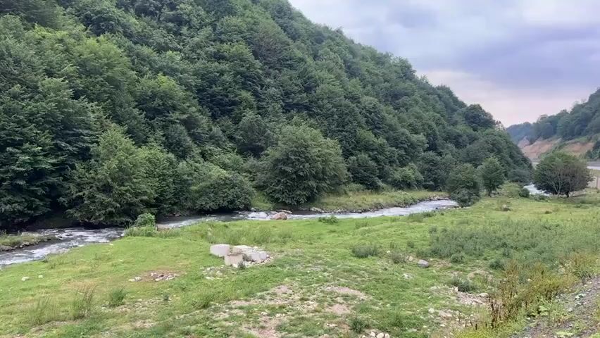 A drone's eye view of a winding river flowing through a lush green valley on an overcast day. The river is fast-flowing with white water and rocks visible. The valley slopes are covered in dense green trees. A small grassy area borders the river, with some concrete debris visible. A road is partially visible in the distance on the right side of the frame. The sky is cloudy and dull.