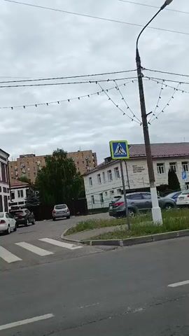 This is a video recording taken from a moving vehicle in Orekhovo-Zuyevo, Russia. The scene depicts a street with several buildings, cars, and utility poles. The sky is overcast, suggesting a cloudy day, possibly in the late morning or afternoon.

The primary subjects are the urban environment and the infrastructure within it. We see a white pedestrian crossing marked on the asphalt road, indicating a controlled intersection. Several cars are parked along the street and on a lot next to a building. These include a white sedan, a dark SUV, a silver SUV, and a dark grey sedan. Overhead, numerous wires crisscross the sky, likely for electricity and telecommunications. A lamppost with decorative string lights is visible on the right side of the frame.

One building appears to be an older, multi-story structure with a red tiled roof and white-framed windows. Another building has a more modern facade with signs. A prominent sign on the rightmost building reads "OREKHOVO-ZUYEVSKY MUSEUM OF LOCAL HISTORY" and another sign below it indicates the years "1945-2025". There is also a sign that says "O FIX".

Traffic signs are visible, including a blue sign with arrows indicating directions for left, straight, and right turns. A yellow and blue pedestrian crossing sign is also present. The overall atmosphere is that of a typical day in a moderately sized Russian city, with no overt signs of unusual activity or strong emotions. The setting appears to be a commercial or mixed-use area.