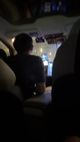 The video shows the interior of a car, with the focus on the driver's seat from a back passenger perspective. The driver, a man wearing a dark t-shirt, is facing forward. Several screens are visible on the dashboard, including a large central display and a phone mounted near the steering wheel. The car is moving, as indicated by the blurred lights visible through the windshield. The interior of the car is illuminated, and the seats appear to be light-colored. A portion of the headrest of the front passenger seat is also visible. The car has a sunroof.