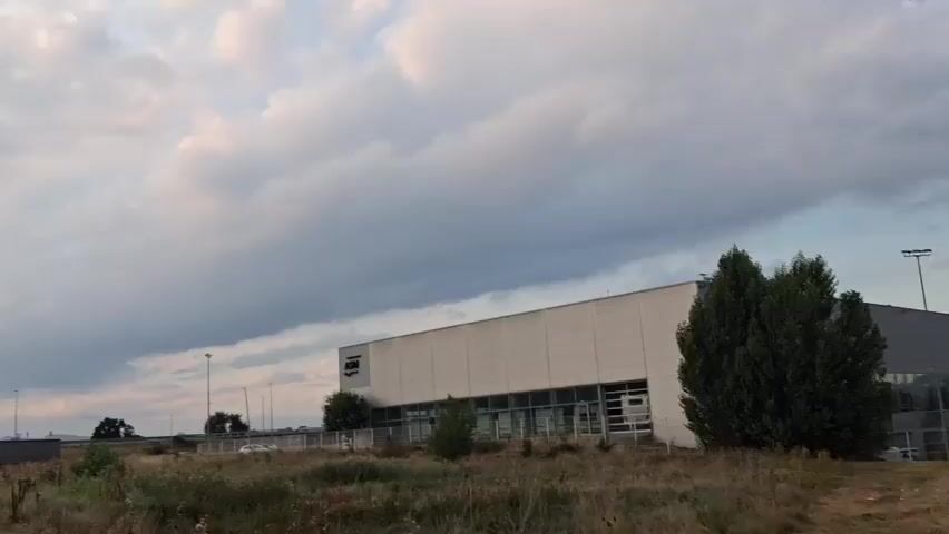 The video begins with a wide shot of an industrial building with a white facade and a row of glass windows. The building has a logo visible on its upper left side. In front of the building, there is overgrown vegetation and a few trees. Beyond the building, a few parked trucks and streetlights are visible. The sky is filled with dramatic, cloudy formations, with sunlight peeking through in some areas. As the video progresses, the camera pans across the sky, focusing on the clouds and the sunlight. The scene appears to be an outdoor setting, possibly an industrial park or a commercial area, with a backdrop of distant hills. The lighting suggests it is either early morning or late afternoon. No people are visible in the video.