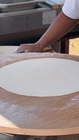 A close-up shot shows a large, thin, oval-shaped piece of white dough spread across a round wooden surface dusted with flour. A hand holding a wooden paddle, with a partially cooked flatbread on it, moves into the frame from the top right and places the flatbread onto the dough. The flatbread is round and appears to be made of a lighter-colored dough with darker browned spots. After placing the flatbread, the hand holding the paddle withdraws from the frame. The background shows a stainless steel counter with a sink and some equipment, suggesting a kitchen or food preparation area. The lighting is bright, casting a shadow on the wooden surface.