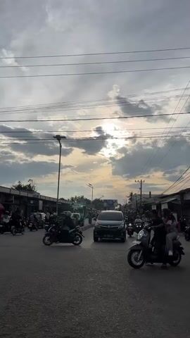 The video shows a street scene at dusk. A few cars and numerous motorcycles are on the road, indicating moderate traffic. The sky is cloudy with the sun peeking through the clouds, casting a soft light. Several buildings line the sides of the street. People are seen riding on motorcycles, with some wearing helmets and headscarves. The overall atmosphere is calm and depicts a typical evening commute in a populated area.