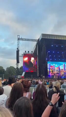 The video shows a large LED screen at an outdoor concert venue. A man wearing a red baseball cap is shown on the screen, his face illuminated by the setting sun. The screen is framed by a red, circular graphic with glowing lines. Below the screen, a crowd of people are visible, facing the stage. The sky is overcast. The concert setup includes scaffolding and speakers.