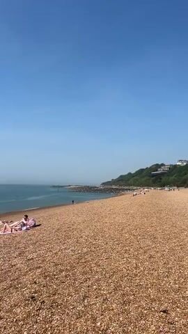 The video presents a static shot of a beach under a clear blue sky. The beach is composed of small, brown pebbles, stretching from the foreground to the ocean. In the foreground, two people are lying on blankets, seemingly sunbathing. Further along the beach, several other individuals can be seen in the distance, some appearing to be relaxing on the shore. The ocean, a calm expanse of blue-gray water, meets the horizon under a hazy sky. A breakwater extends into the ocean. Vegetation covers a hill in the background. The overall ambiance of the scene suggests a warm, sunny day at the beach.