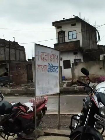The video shows a street scene. In the foreground, there is a wall with text written on it in Hindi. The text appears to be the name of a medical store. To the right of the wall, there is a building with a doorway and some stairs. In the background, there are more buildings, some of which appear to be made of corrugated metal. There is also a tree and some people walking around. A motorcycle is parked in the foreground. The overall impression is of a busy street in a developing country.