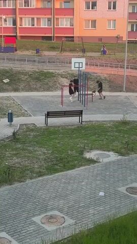 Two young males are in an outdoor park area. One male is wearing a black t-shirt and black shorts and is using a horizontal bar for exercise, with his feet touching the ground. The second male is wearing a black t-shirt and black shorts and is standing next to him, looking at him. A third male is wearing a black t-shirt and patterned shorts and is standing near a red soccer goal frame with a white net. There is a basketball hoop mounted on a blue pole. The ground is paved with gray tiles and there is grass in the foreground and background. A wooden bench is situated in the midground. In the background, there is a multi-story building and playground equipment including swings and a slide. The overall lighting suggests it is late afternoon or early evening.