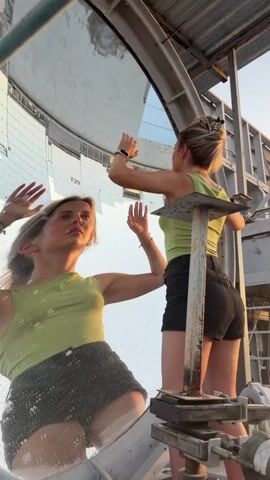 A woman with blonde hair tied up in a bun, wearing a light green tank top and black shorts, stands in front of a large, reflective surface, which appears to be a parabolic mirror. Her reflection is visible in the mirror, and she is interacting with it. In the foreground, a metal support structure is visible, with rusty metal components. The background suggests an outdoor industrial or technological setting with some building structures and a light sky. The woman is shown from different angles throughout the video, sometimes facing the mirror, sometimes looking away, and her hands are raised and moving, suggesting she is posing or playing with her reflection. The lighting suggests it is daytime, possibly late afternoon due to the warm tones.