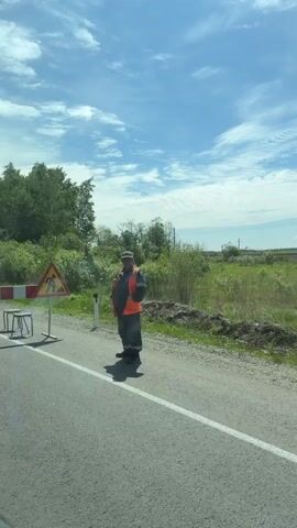 A video captures a roadside scene with a blue sky and scattered clouds. A worker in an orange vest and gray pants stands on the right side of a paved road, facing left, holding a phone. A road construction sign, depicting a shovel and pickaxe, is positioned to the left of the worker. Further left, a barricade with red and white stripes is set up, with two stools behind it. Dense green foliage and trees line the background. The scene appears to be a temporary road closure or construction zone.