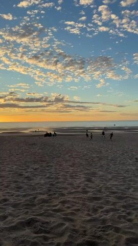 A vertical, wide shot shows a beach scene at sunset. The sky is a vibrant blue with scattered, puffy white clouds tinged with orange and yellow from the setting sun. The horizon displays a gradient of warm colors transitioning from yellow to orange, with the ocean appearing as a calmer, darker band. In the foreground, the sand is textured with ripples from the tide. Scattered across the mid-ground of the beach, several small figures are visible. To the left, a group of people are sitting or standing near the water's edge. Further to the right, a few individuals are walking or playing, and at least one appears to be a dog. The overall impression is one of a serene, natural environment with human activity.
