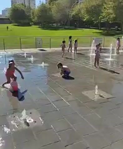 A group of children are playing in a splash pad on a sunny day. There are fountains spraying water from the ground, and the children are running through them and getting wet. A few adults are present, watching the children. In the background, there is a grassy park area with trees and buildings. The splash pad is made of gray stone tiles. One child is wearing a pink swimsuit and a hat. Another child is crawling on the wet tiles. A woman is walking through the splash pad, and a man is standing to the side, holding a phone.