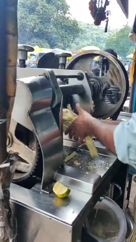 The video shows a person operating a sugarcane juice machine on a street. The machine is made of stainless steel and has large gears. Sugarcane stalks are fed into the machine, and juice is extracted. A half-cut lemon is visible on the machine. In the background, a bus and a motorcycle are on the road, and there are trees and some market stalls. The overall environment appears to be a busy street market.