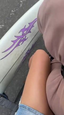 The video begins with a close-up of a person's legs and part of a surfboard with purple tribal decals. The camera then pans to the back of another person wearing a pink t-shirt and a helmet, suggesting they are riding a motorcycle.

The scene transitions to a wider shot of a coastal road in Denuwala, Sri Lanka. Two men are standing on a bridge overlooking the ocean. The sky is blue with scattered clouds, indicating it's daytime with fair weather. The ocean appears calm with gentle waves. The bridge has a white railing with black and white checkered patterns on the road. There is a streetlight on the left side of the bridge and trees in the background. A bicycle is parked on the right side of the bridge. The overall mood seems relaxed and scenic, typical of a coastal environment. No text is visible in the video.