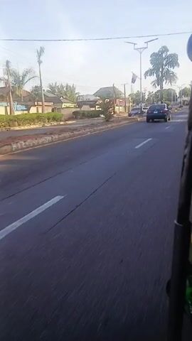 The video shows a view from a moving vehicle driving on a paved road.  The road has a white dividing line.  Buildings and houses are visible on both sides of the street behind a concrete curb. Trees are planted intermittently along the roadside. Cars are parked and moving along the street. Streetlights are present. The sky is light blue with some clouds. The footage is a bit shaky due to the motion of the vehicle.