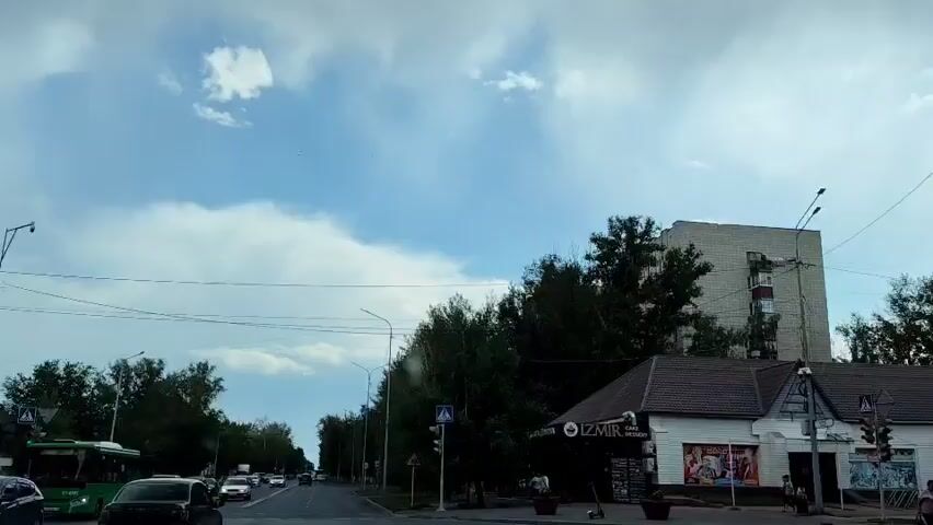 The video shows a street scene with a blue sky and white clouds. There are several buildings on the right side of the street, some with tiled roofs. Trees line the street. There are cars and pedestrians on the street and sidewalk. A crosswalk with a traffic light is visible. One building has a sign that says "IZMIR". A tall, light-colored building is visible behind the trees. The video appears to be filmed from a moving vehicle.