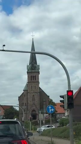 The video begins with a wide shot of a church tower with a green spire and a clock. A traffic light is visible in the foreground, showing a red light. The camera then zooms in on the church tower. The scene appears to be a street in a town or city, with buildings and trees in the background. The sky is cloudy.