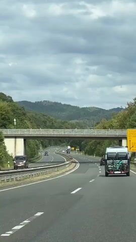 The video shows a view from inside a vehicle traveling on a highway. On the right side of the highway, a camper van with bicycles mounted on the back is visible. Other vehicles, including cars, are also on the highway. An overpass with a white railing crosses the highway. The highway is surrounded by lush green trees and hills in the background under a cloudy sky. The road has multiple lanes and markings.