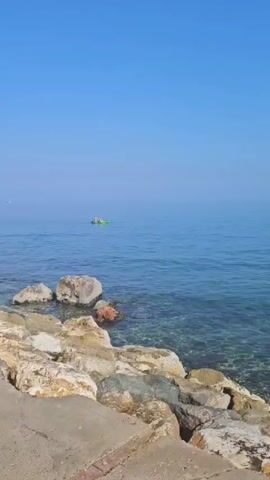A long shot shows a body of water with a clear sky above. The water is a deep blue and extends to the horizon. In the foreground, a rocky shore is visible, with large boulders and some smaller rocks lining the edge of the water. On the water, a small, green inflatable boat with two people in it is paddling away from the shore. The camera follows the boat as it moves further into the distance. The sounds of conversation can be heard in the background.