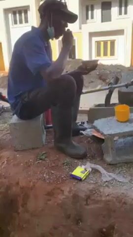 A man wearing a blue polo shirt, dark trousers, black boots, and a dark baseball cap sits on a concrete block. He also wears a face mask. The man eats from a small bowl. To his right is a concrete block structure supporting a yellow cup. A box of matches and plastic trash are on the ground next to the concrete blocks. A yard brush is leaning against one of the blocks. Behind the man is a building with white walls and multiple windows. The area around him is an unfinished construction site.