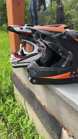 The video opens with a close-up view of two motocross helmets resting on a wooden surface. The helmet on the left is white with red and black accents and white goggles. The helmet on the right is black with orange accents and a clear visor. The scene transitions to a wider shot, revealing a grassy area with yellow wildflowers. Several pairs of footwear, including boots, gloves, and flip-flops, are laid out on the wooden surface. In the background, clothes are hanging on a makeshift clothesline constructed from wooden posts. The overall environment appears to be outdoors, possibly in a rural or backyard setting, with lush green vegetation and trees.